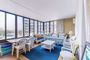 A modern living room with large windows, a sofa set, white wicker furniture, and a blue rug. - Beachfront condo with balcony, W/D, & outdoor pool (Ocean City)