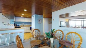 Uma cozinha moderna com armários brancos, uma ilha central com banquetas e um espaço de refeições com mesa de vidro e cadeiras de vime.