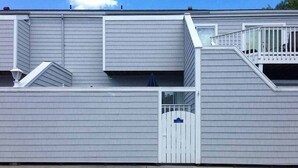 A two-story building with a garage door, a small balcony, and a white fence. - Condo 3 Blocks from the Beach w/ Sunny Patio, Central AC, WiFi & Shared Pool (Ocean City)