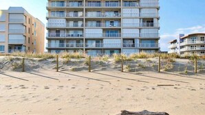 Un hotel de varios pisos con balcones, situado cerca de una playa de arena con dunas.