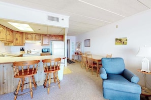 A compact kitchen with wooden cabinets, a central island with stools, a blue armchair, and a dining table. - Well-Equipped Family Beachfront Condo w/Balcony, Views, Washer/Dryer, A/C, WiFi (Ocean City)