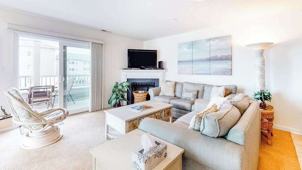 A living room with a sofa, armchair, fireplace, TV, and a balcony with a view. - Waterfront condo with pool, balcony, fireplace, central AC, & washer/dryer (Ocean City)