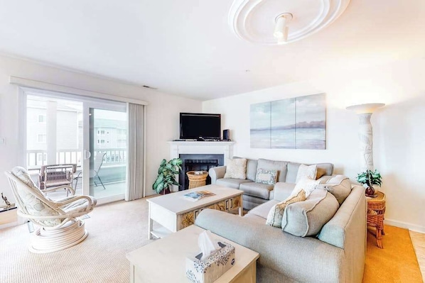 A living room with a sofa, armchair, fireplace, TV, and a balcony with a view. - Waterfront condo with pool, balcony, fireplace, central AC, & washer/dryer (Ocean City)