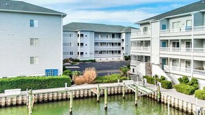 A waterfront apartment complex with multiple balconies, a parking lot, and a small bridge over the water. - Waterfront condo with pool, balcony, fireplace, central AC, & washer/dryer (Ocean City)