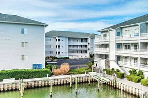 A waterfront apartment complex with multiple balconies, a parking lot, and a small bridge over the water. - Waterfront condo with pool, balcony, fireplace, central AC, & washer/dryer (Ocean City)