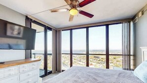 A bedroom with a bed, a dresser, a ceiling fan, and large windows offering a city view. - Cozy beachfront condo with balcony views, pools & game room (Ocean City)