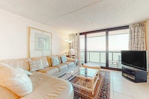 Ein Wohnzimmer mit einer Couch, einem Couchtisch, einem Fernseher und einem großen Fenster mit Vorhängen.