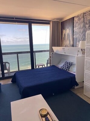 A hotel room with a large window, a bed, a desk, and a chair. - Beachfront studio with balcony, washer/dryer, pools, & gym (Ocean City)