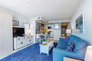 A modern living room with a flat-screen TV, a sofa, a coffee table, and a dining area with a table and chairs. - Beachfront studio with balcony, washer/dryer, pools, & gym (Ocean City)