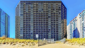 A high-rise building with a grid of windows, surrounded by sand dunes and other buildings. - Beachfront studio with balcony, washer/dryer, pools, & gym (Ocean City)