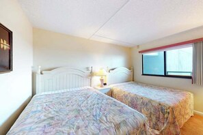 Et soveværelse med to senge, et vindue med gardiner, et sengebord med en lampe og et billede i ramme på væggen.