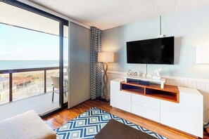 Ein modernes Wohnzimmer mit großem Fernseher, einer Couch, einem gemusterten Teppich und Blick aufs Meer.