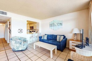 A living room with a blue sofa, white coffee table, and a dining area with chairs. - Fountainhead Towers 1206 (Ocean City)