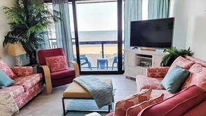 Olohuone, jossa on televisio, sohvia, punainen nojatuoli, sohvapöytä ja suuret ikkunat, joista avautuu näkymä rannalle.