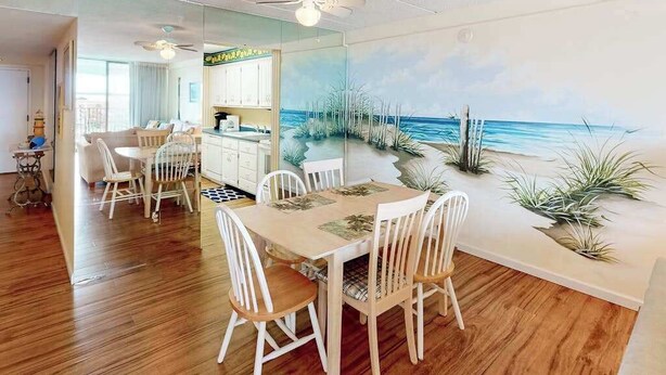 Un coin repas avec une table et des chaises en bois, un ventilateur de plafond et une fresque murale représentant une plage.