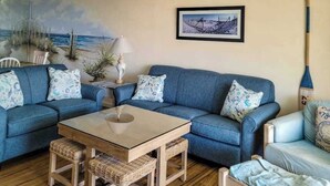 Un salon avec un canapé bleu, une table basse en bois, des fauteuils en osier et un tableau représentant un paysage enneigé.