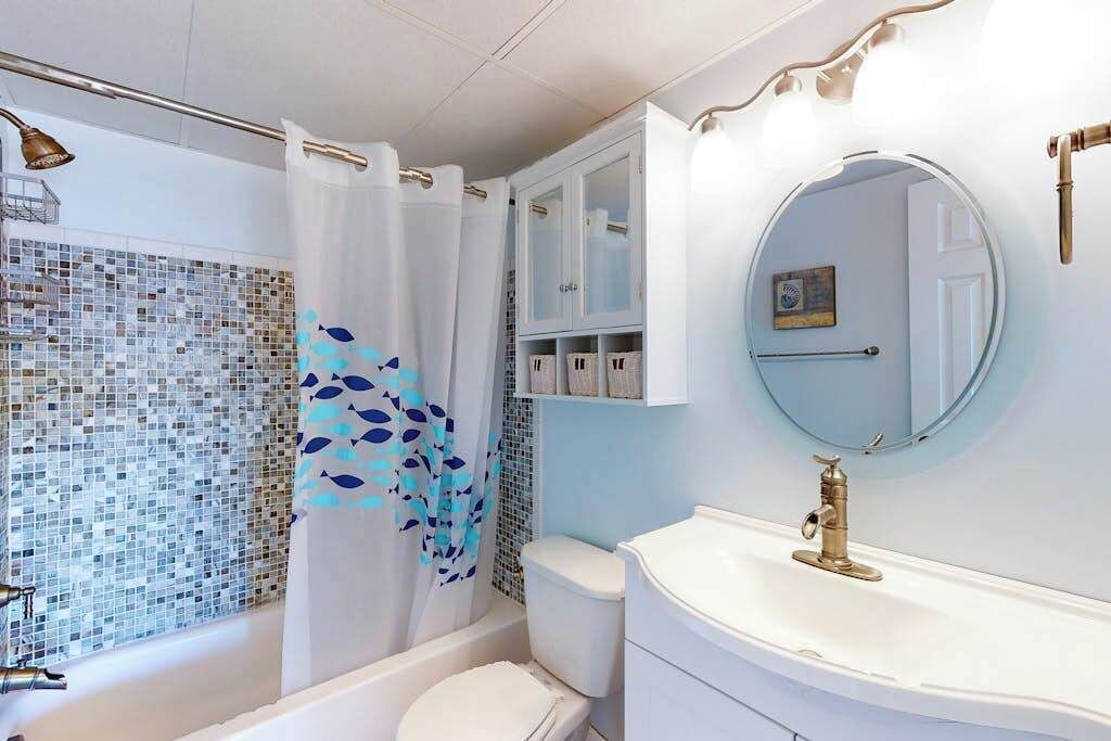 Une salle de bain avec un mur recouvert de mosaïques, un rideau de douche orné d’un motif de poissons, des toilettes blanches, un meuble-lavabo avec un robinet doré et un miroir.