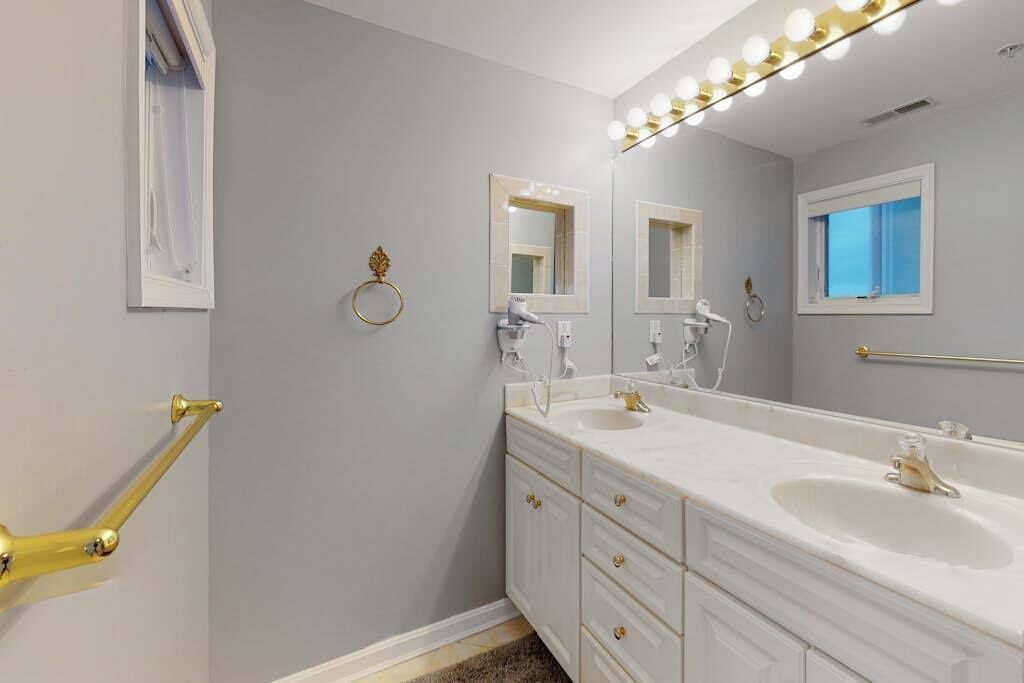 A bathroom with two sinks, a large mirror with lights, a towel rack, and a window.