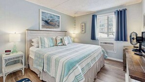 Een slaapkamer met een bed, nachtkastje, lamp, raam met gordijnen en een ventilator aan de muur.