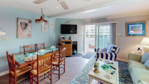 A living room with a dining table, chairs, a sofa, a coffee table, a ceiling fan, a TV, and a view through a sliding door. - Cinnamon Teal 10 (Ocean City)