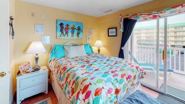 A bedroom with a bed, two bedside lamps, a nightstand, a painting, and a view of the outside. - Cinnamon Teal 10 (Ocean City)