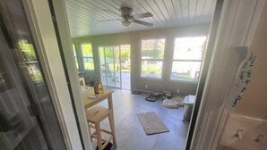A room with a ceiling fan, a dining table with chairs, a pet area with a dog bed and bowls, and a large window with a view of the outdoors.