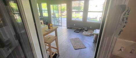 A room with a ceiling fan, a dining table with chairs, a pet area with a dog bed and bowls, and a large window with a view of the outdoors.