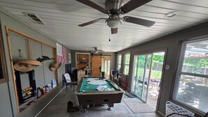 A pool table in a room with a ceiling fan, a window with a view of the outdoors, and a door leading to another area.