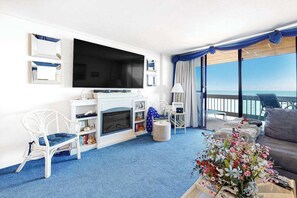 A hotel room with a large TV, a fireplace, a sofa, and a balcony with a view. - Attractive oceanfront condo with sunset views, two balconies, pool & Central AC (Ocean City)