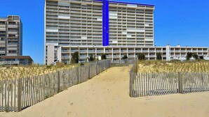 A multi-story hotel with a central blue vertical feature, a sandy pathway leading to the entrance, and a wooden fence on both sides. - Carousel 1707 (Ocean City)