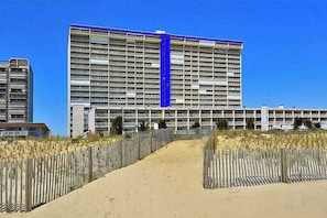 A large hotel with multiple floors, a prominent blue vertical stripe, and a sandy beach area in the foreground. - Beachfront Condo with balcony, views, shared pools, gym, & sauna (Ocean City)