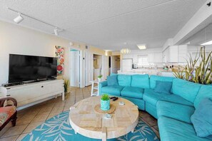 A living room with a television, a sofa, a coffee table, and a kitchen in the background.