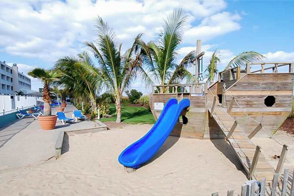 Una zona sulla spiaggia con uno scivolo blu, palme e poltrone relax.
