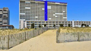 A large hotel with multiple floors, a sandy pathway leading to the entrance, and a wooden fence on either side.