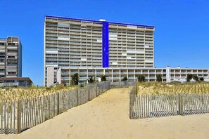 A large hotel with multiple floors, a sandy pathway leading to the entrance, and a wooden fence on either side.