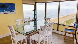 A dining area with a glass table and white chairs, a telescope, and a painting of a beach scene. - Beachfront Condo w/Private Balcony, Telescope, Shared Pool, A/C, Free WiFi (Ocean City)