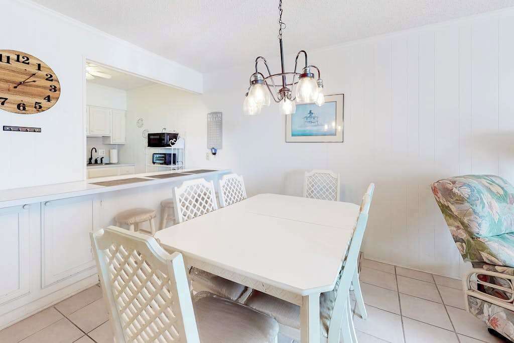 Comedor con mesa y sillas blancas, cocina al fondo y una araña.