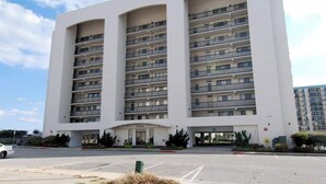 A multi-story hotel with balconies, a central entrance, and a parking lot. - Camelot 203B (Ocean City)