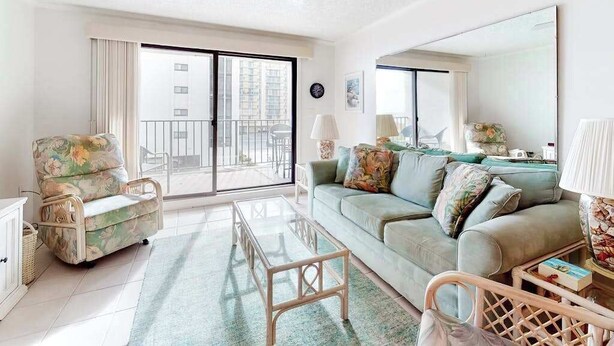 A living room with a sofa, armchair, coffee table, and a large window with a view of buildings. - Camelot 203B (Ocean City)