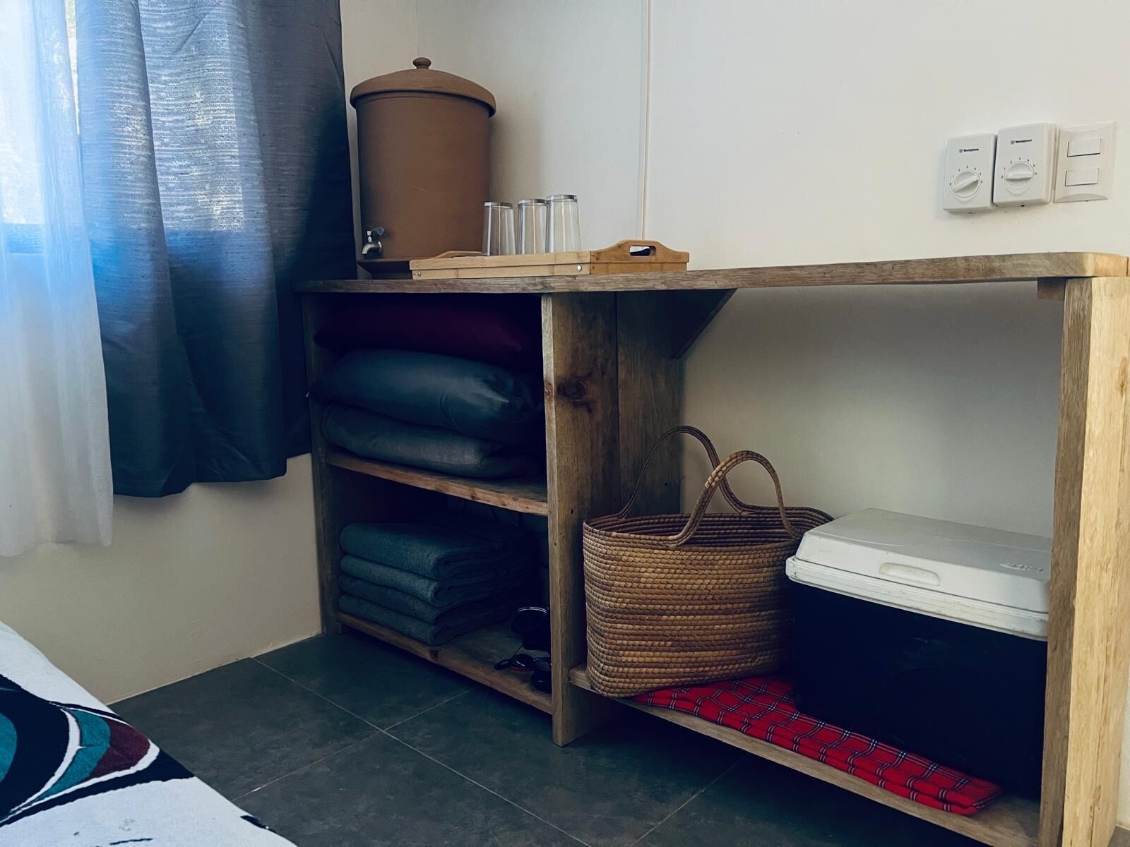 Una habitación con un escritorio de madera, estantes con ropa doblada, una cesta de mimbre y un cubo de basura.