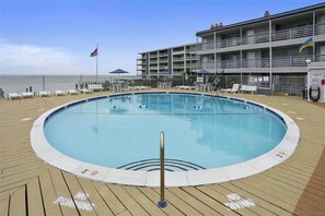 A pool area with lounge chairs, a flag, and a building with balconies. - Bradley On The Bay 130G (Ocean City)