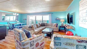 A living room with a sofa, coffee table, and a dining area with a table and chairs. - Assateague House 205 (Ocean City)