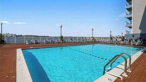 A swimming pool with a wooden deck, lounge chairs, and a building with balconies. - Assateague House 205 (Ocean City)