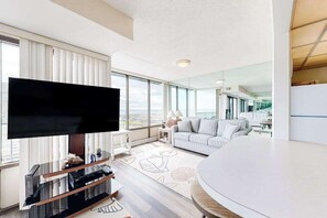 A modern living room with a flat-screen TV, a sofa, a dining table, and large windows offering a city view. - Bright coastal condo with pool access, side views of the ocean, balcony & W/D (Ocean City)