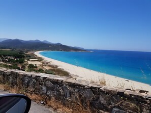 A coastal landscape with a beach, clear blue sea, and distant mountains.