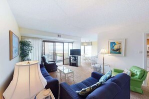 A living room with a sofa, a TV, a lamp, a green chair, and a painting on the wall. - Beachfront Condo w/Private Balcony, Shared Pool, Ocean Views, Free WiFi, A/C (Ocean City)