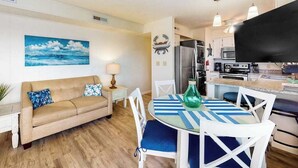 A living room with a sofa, a dining table with chairs, a kitchen area with appliances, and a painting on the wall.