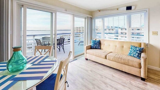 A room with a sofa, a table with a striped tablecloth, a vase, and a view of the sea through large windows.