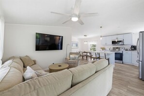 A modern living room with a sofa, a television, a ceiling fan, and a kitchen area with cabinets and appliances. - Cozy 2-bedroom mobile home in cool Fort Myers Beach with AC (Fort Myers Beach)