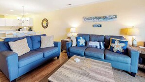 Ein Wohnzimmer mit blauem Sofagarnitur, einem Couchtisch und einer Küche im Hintergrund.